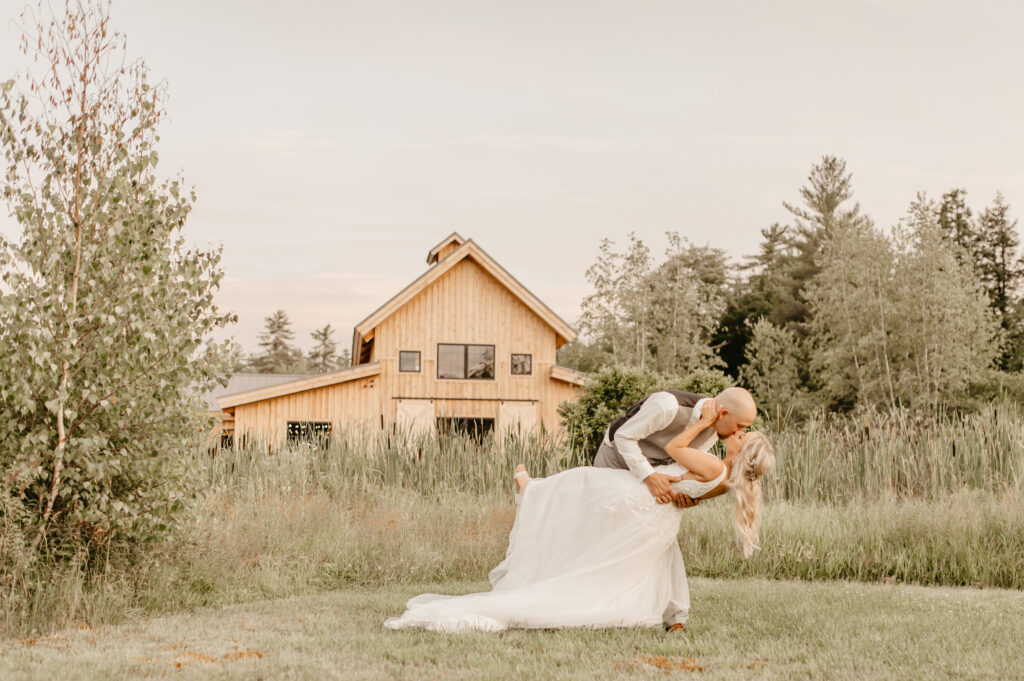 At their wedding, a groom playfully dips his bride for a kiss in front of a rustic barn surrounded by lush greenery. The bride's flowing white dress and the groom's vest and trousers enhance the serene, romantic scene set against a soft, natural backdrop.