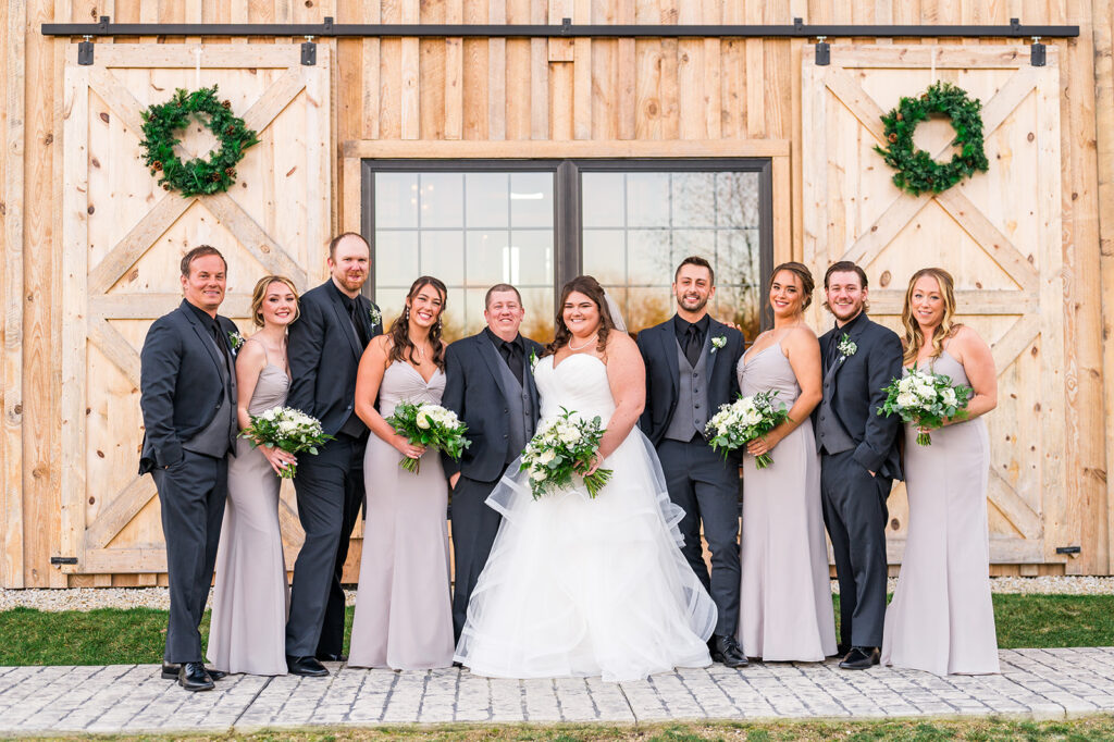 A bride in a white gown and a groom stand with eight other people in formal attire in front of a rustic wooden wedding venue adorned with wreaths. The bridesmaids wear matching dresses and hold bouquets, while the groomsmen don dark suits. Everyone is smiling.