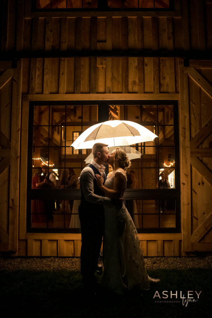 A couple in wedding attire embraces under a lit umbrella at night, capturing the essence of a perfect wedding moment. In front of a wooden building with large windows, warm indoor lights illuminate the scene, creating a romantic atmosphere.