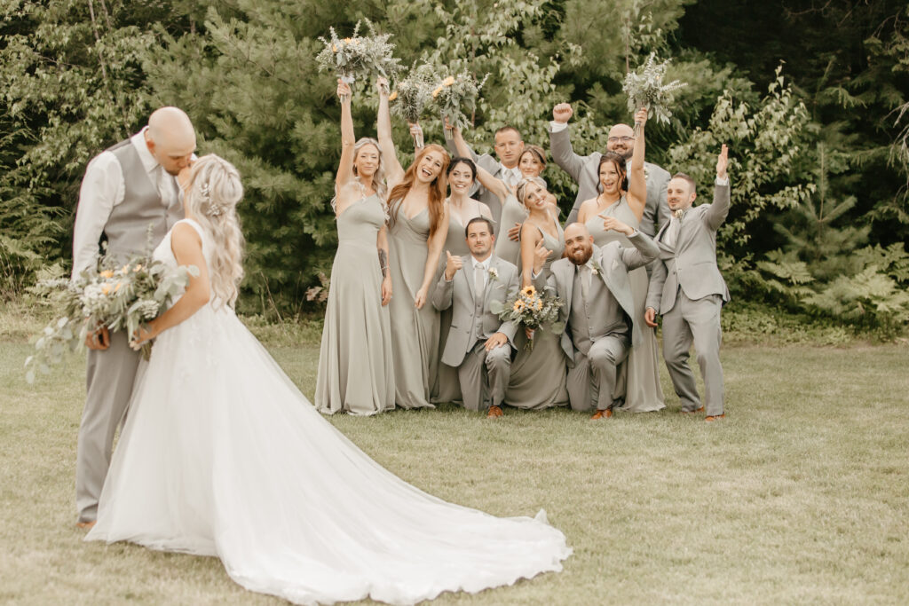 A bride and groom share a wedding kiss in the foreground, while a joyful wedding party in matching gray attire celebrates behind them. The scene is set in a lush, green outdoor area with trees. Bridesmaids raise bouquets, and groomsmen cheer enthusiastically.