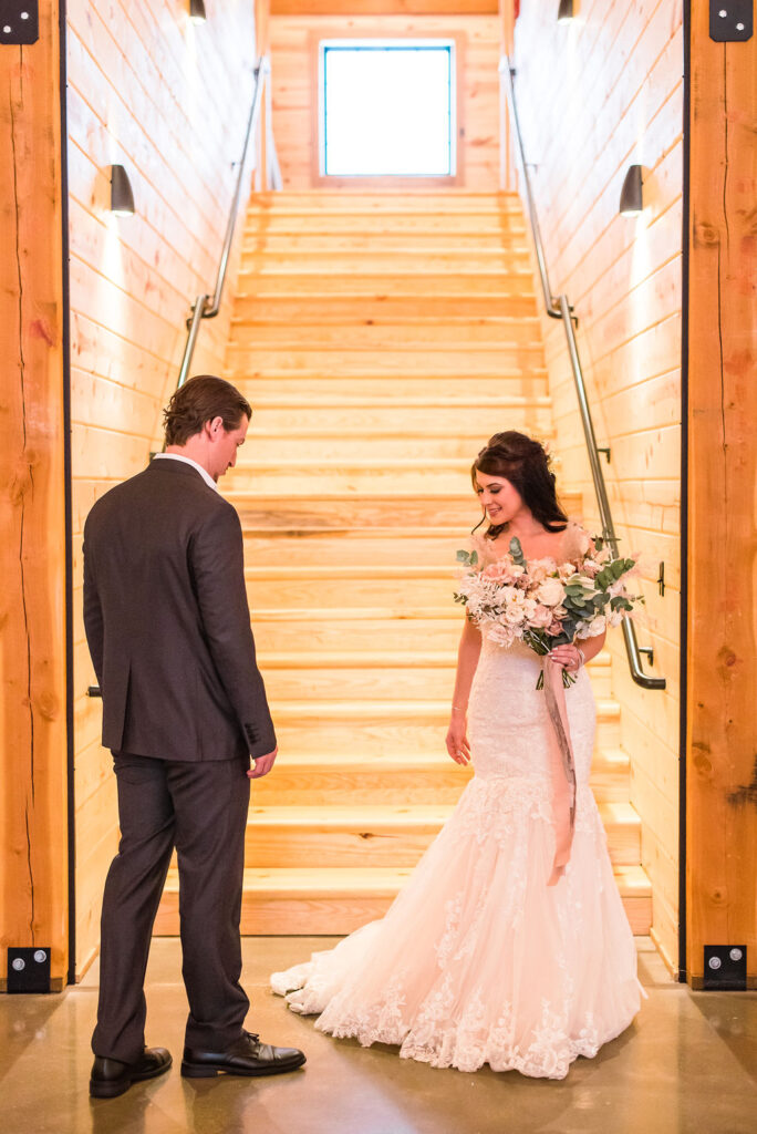 A bride in a white lace gown holds a bouquet and smiles at the groom in a dark suit. They stand together before a bright wooden staircase, embodying the warm, rustic charm of their wedding day.