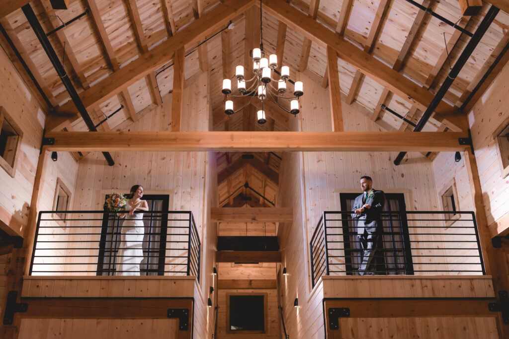 The bride and groom stand on separate balconies in a rustic wooden barn, the perfect setting for their wedding. A chandelier hangs overhead as the bride holds a bouquet and the groom looks dapper in his suit, capturing a moment of timeless romance amidst charming wooden beams.