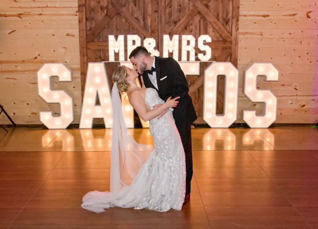 The wedding venue is a dream come true as the bride and groom share a romantic dance on the wooden floor. Behind them, large illuminated letters spell out "Mr & Mrs Santos" against a rustic wooden backdrop, capturing the essence of their special day.