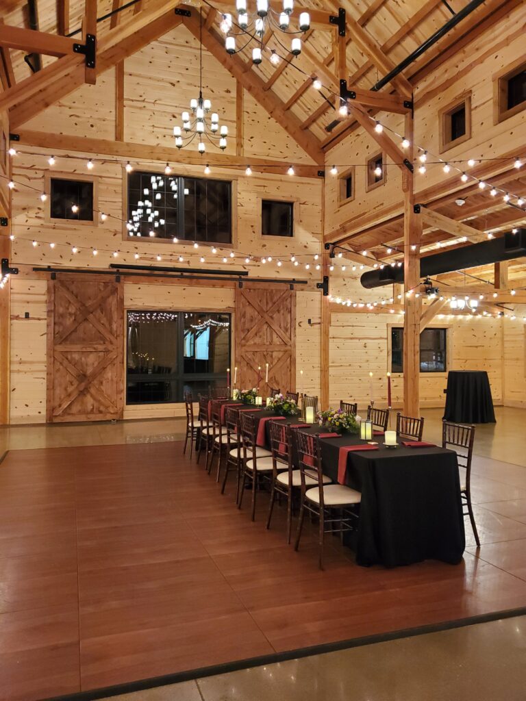 A spacious wooden barn interior with high ceilings and string lights creates the perfect wedding ambiance. A long dining table adorned with a dark tablecloth, chairs, and candlesticks sits at the center. Large barn doors and a softly lit atmosphere complete this rustic, romantic setting.