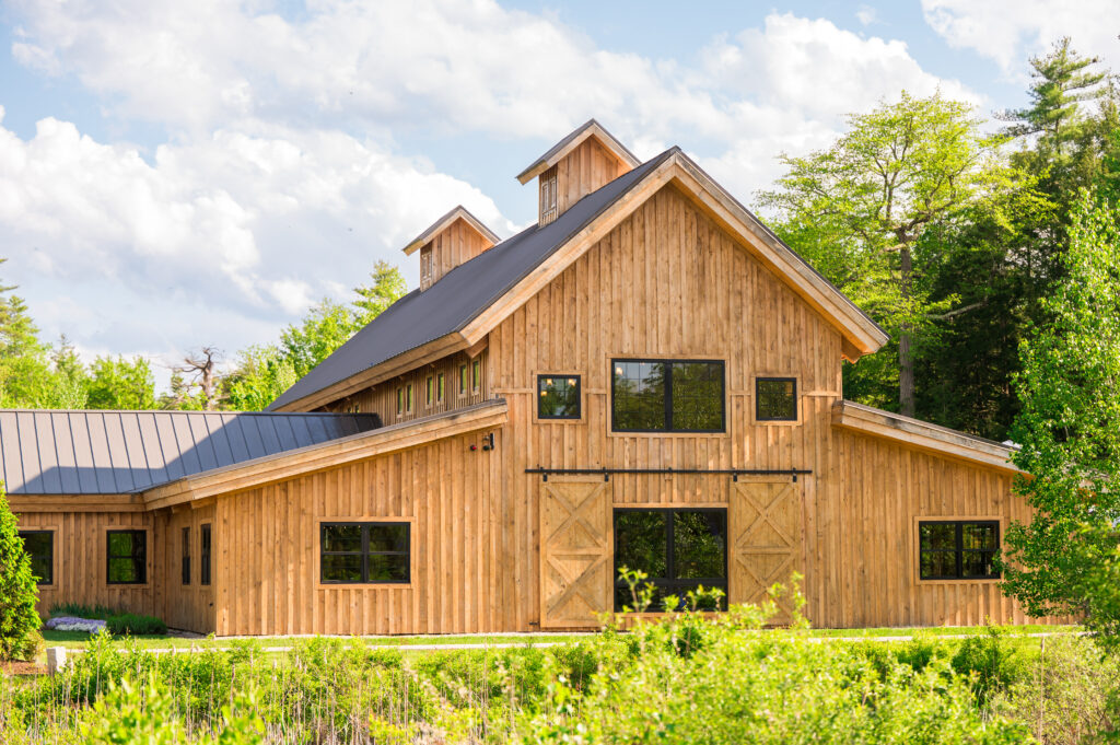 A large wooden barn with a metal roof is surrounded by lush greenery and trees under a partly cloudy blue sky. The barn features large double doors and several windows, giving it a rustic appearance.