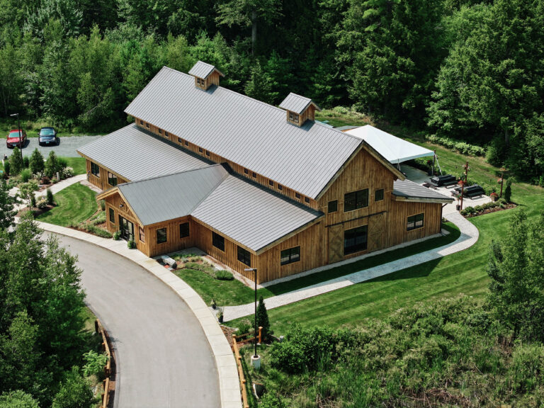 A rustic, large wooden building with a metal roof, surrounded by lush greenery. There are two parked cars near a paved road curving around the structure, and a patio area with seating on one side.