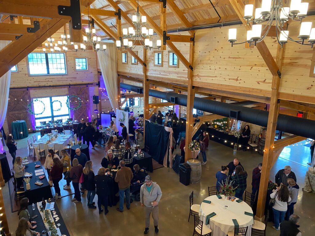 A lively gathering inside a large wooden barn, decorated with lights and drapes. People socialize around tables and displays, while overhead chandeliers illuminate the spacious interior. Natural light filters through large windows.