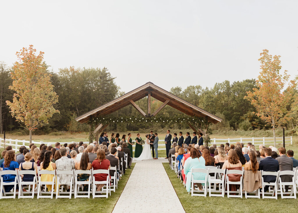 A wedding ceremony is taking place outdoors under a wooden arch with string lights. Guests are seated on either side of a white aisle, surrounded by green lawns and trees with autumn leaves. The officiant stands with the couple at the altar.