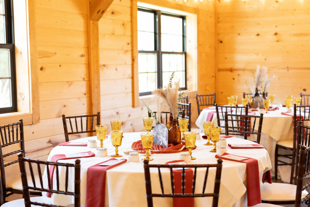 A warmly lit dining area with wooden walls features round tables set with yellow glasses, white plates, and red napkins. Each table has a centerpiece with decorative elements. Brown chairs surround the tables, and sunlight streams through the windows.