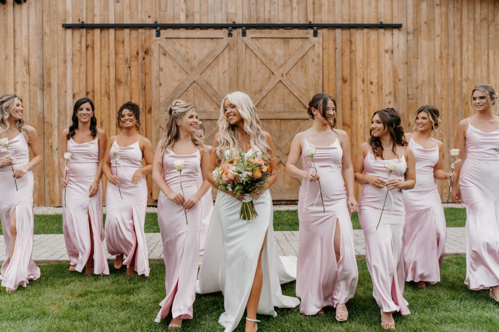A bride in a white gown holds flowers and smiles while standing with bridesmaids in pink dresses. They are outdoors in front of a wooden barn, standing on grass. Each bridesmaid holds a single white rose.