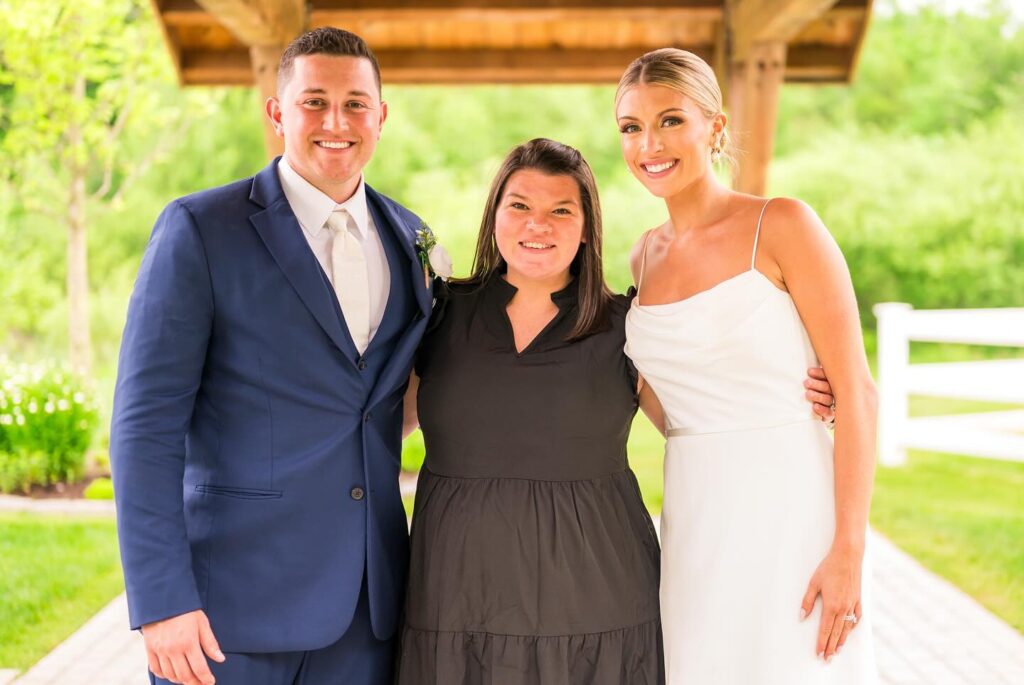 Three people stand smiling outdoors under a wooden structure. The man on the left wears a blue suit and white tie, the woman on the right wears a white dress, and the woman in the middle wears a black dress. Greenery is visible in the background.