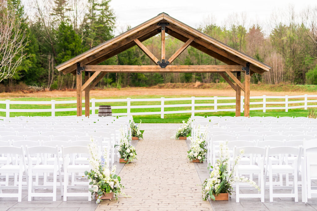 An outdoor wedding setup with rows of white chairs on either side of an aisle decorated with white flowers, facing a wooden arbor, with green grass and trees in the background.