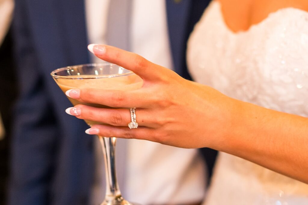A bride in a white strapless dress holds a cocktail glass, showing a diamond engagement ring. A person in a blue suit stands beside her, partially out of focus.