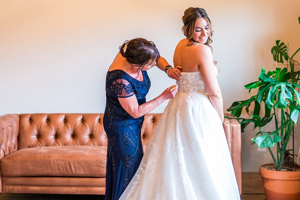 A woman in a navy dress helps a bride in a white wedding gown adjust her dress. They stand in a bright room with a tan leather sofa, a plant, and wooden beams overhead. The bride smiles, looking over her shoulder.