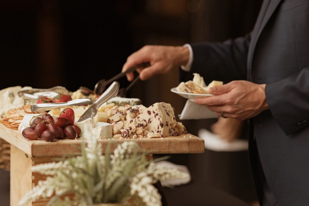 A person in a suit serves cheese and grapes onto a small plate from a wooden cheese board at a buffet; a floral arrangement sits in the foreground.