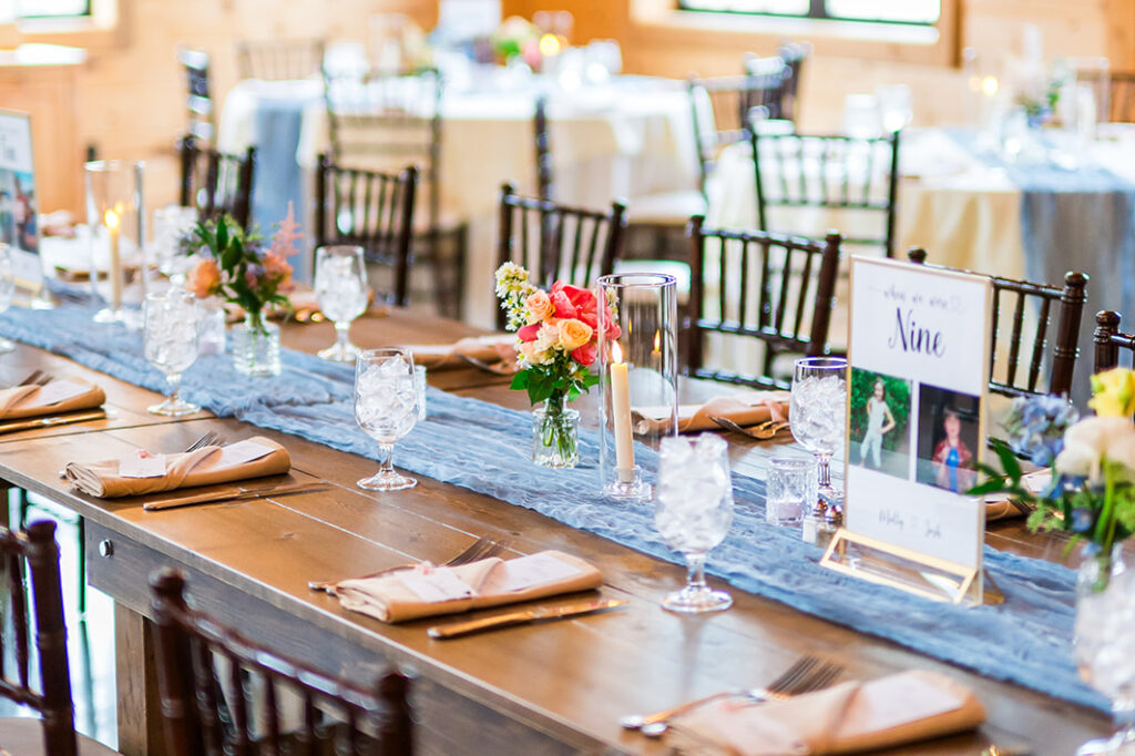 A rustic wedding reception setup with wooden tables, surrounded by brown chairs, floral centerpieces, candles, and table numbers. Soft string lights hang from the wooden ceiling, and natural light enters through large windows.