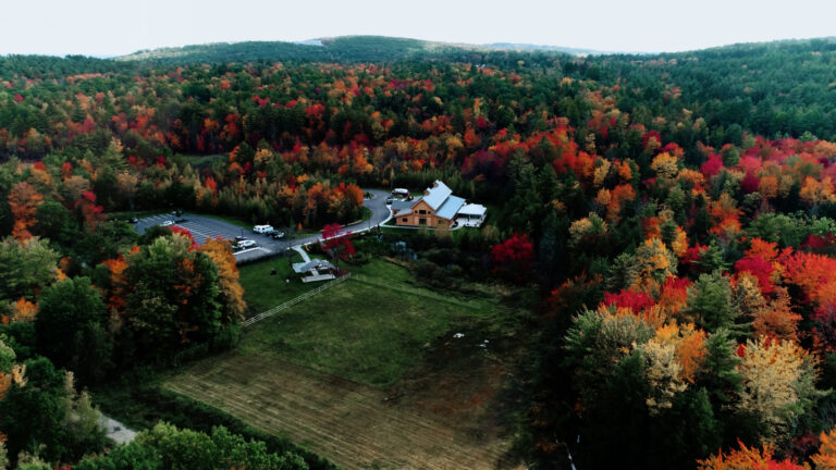 Aerial view of a large cabin surrounded by vibrant autumn trees in red, orange, and yellow hues, with a parking lot and cars nearby, and a grassy field in the foreground. Forested hills extend into the distance.