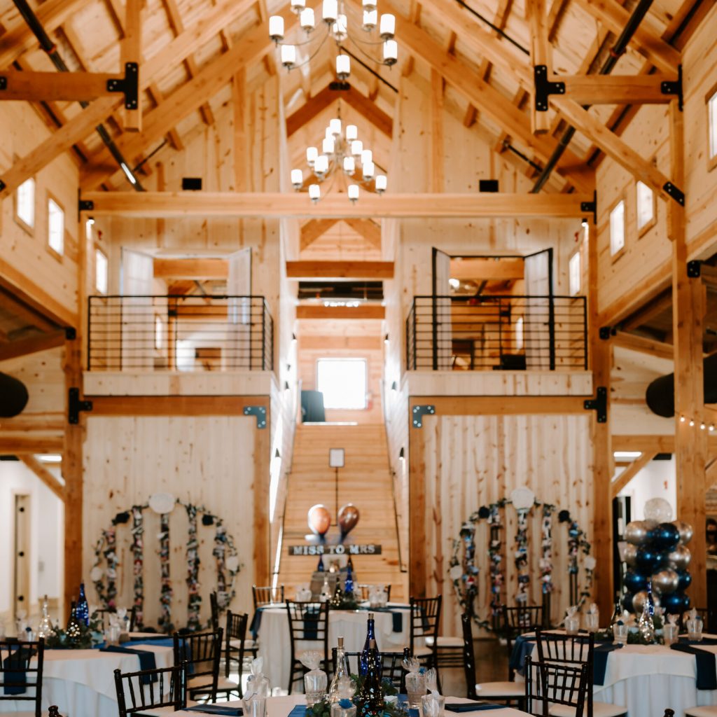 A rustic wedding venue with high wooden ceilings and chandeliers. Round tables are set with white tablecloths, blue napkins, and elegant centerpieces. Decorative floral arrangements adorn the space, creating a charming ambiance.