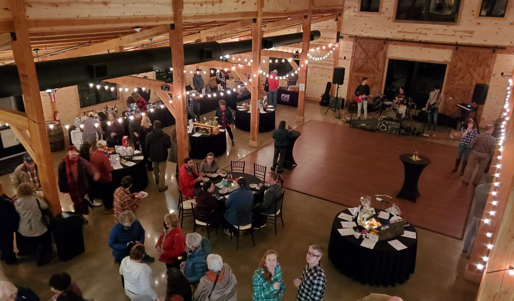 A lively gathering in a rustic wooden hall with string lights. People socialize around tables and at a small stage where a band performs. The atmosphere is warm and inviting, with a mix of standing and seated guests.