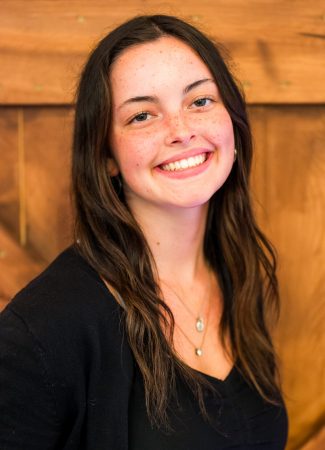 A woman with long brown hair and freckles smiles warmly at the camera. She is wearing a black top with a necklace, standing against a wooden background.